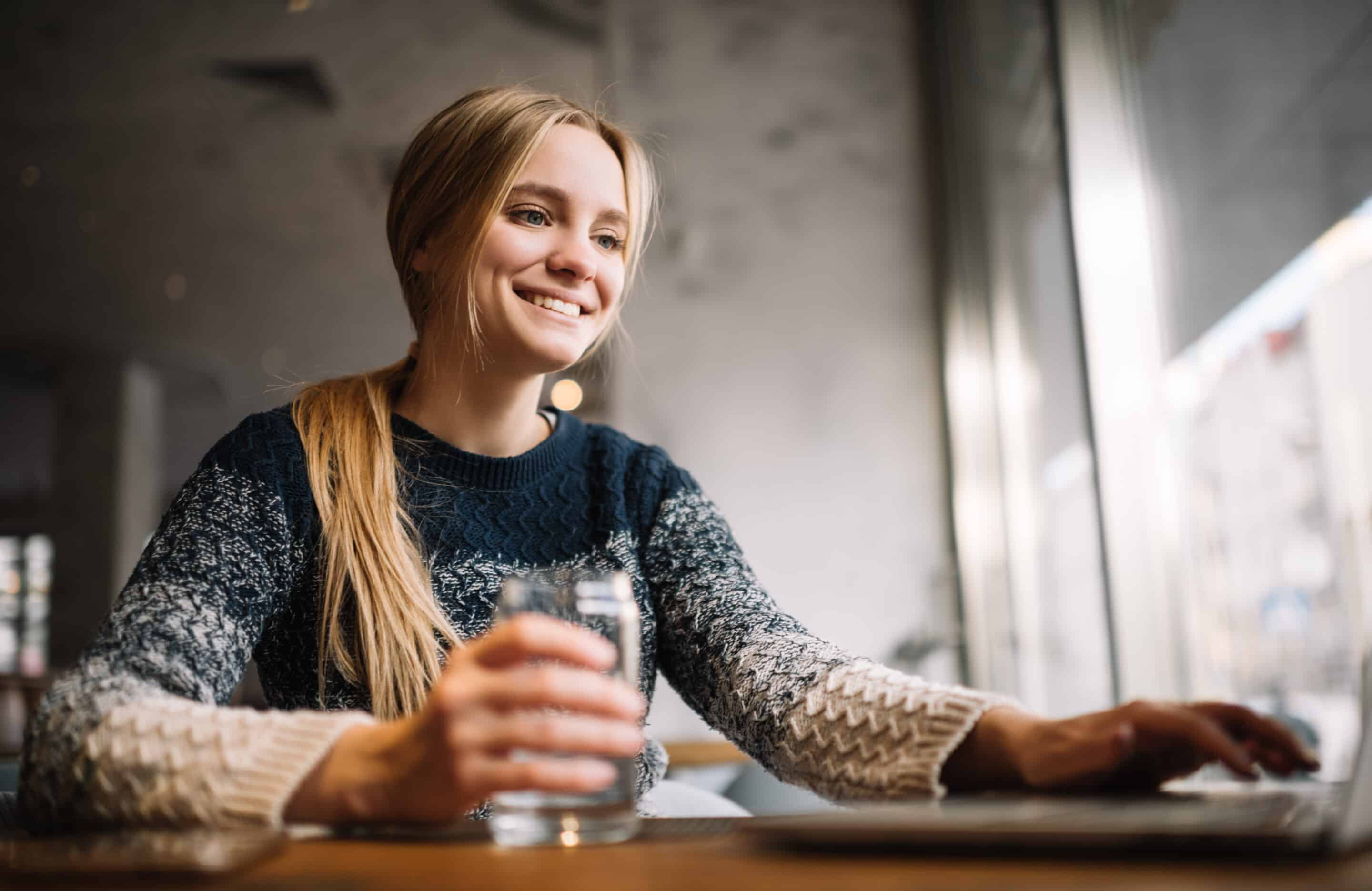 Portrait of young smiling woman sitting in loft cafe using laptop computer and internet, typing on keyboard, researching information. Attractive freelancer copywriter at work. Successful business.